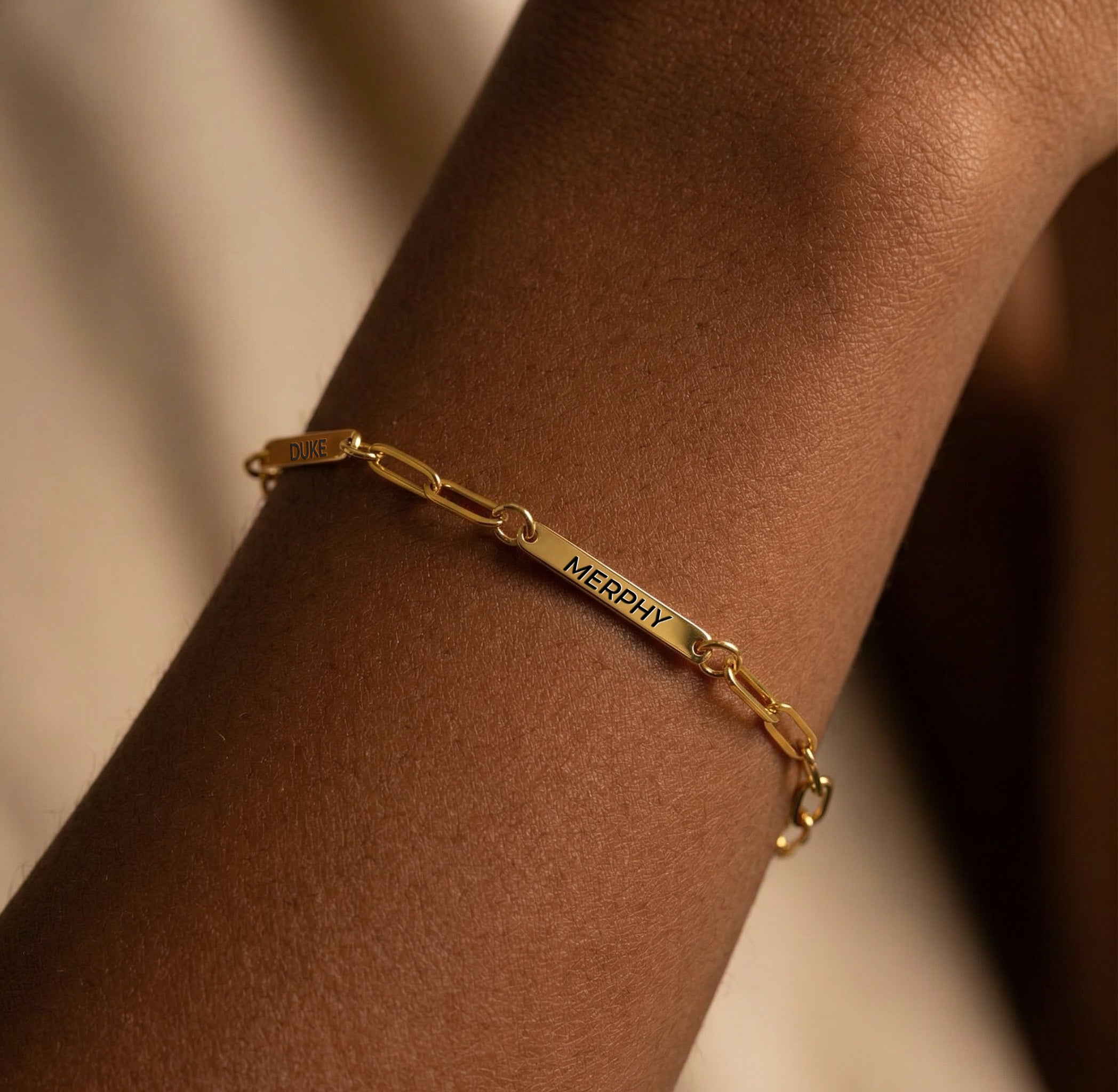 Gold paperclip chain pet name bracelet with engraved name bars worn on a wrist in warm light