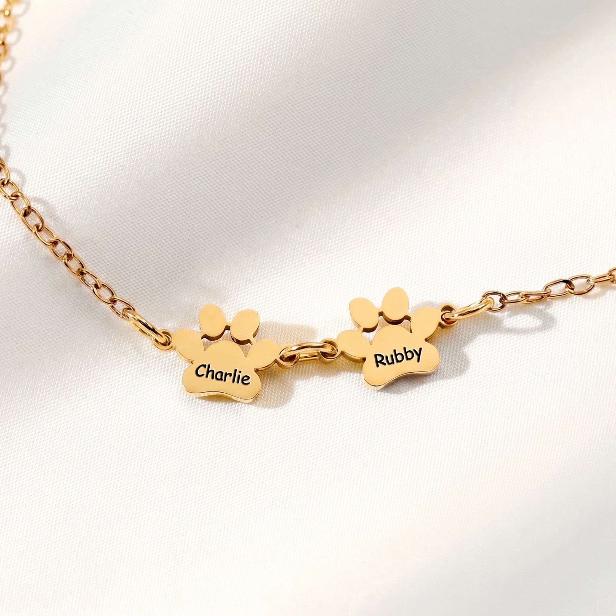 Two gold paw name charms on an adjustable chain bracelet on white satin