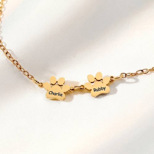 Two gold paw name charms on an adjustable chain bracelet on white satin