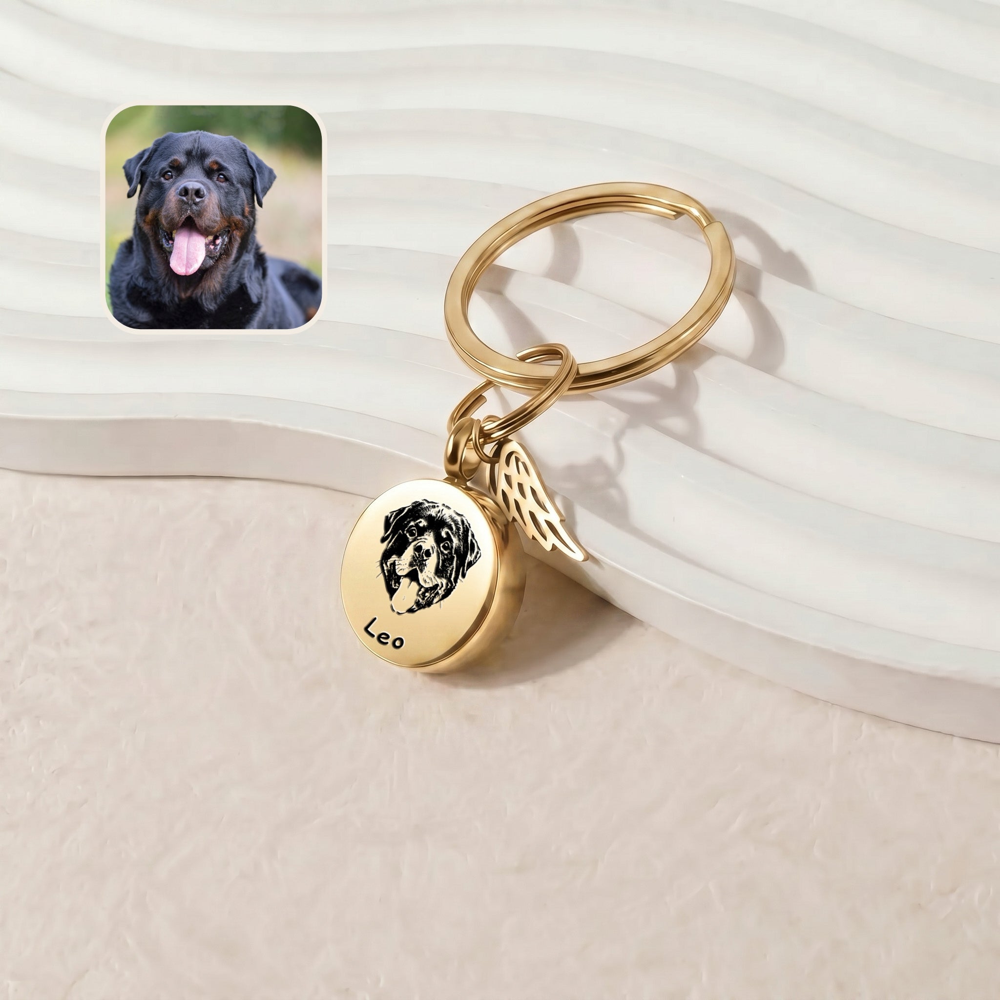 18k gold-plated pet cremation disc keychain &mdash; hand-drawn portrait engraved on a 20mm screw-top disc with permanently-attached angel-wing memorial charm, leaned against a ribbed ceramic tray on a cream plush surface beside a reference dog photo inset