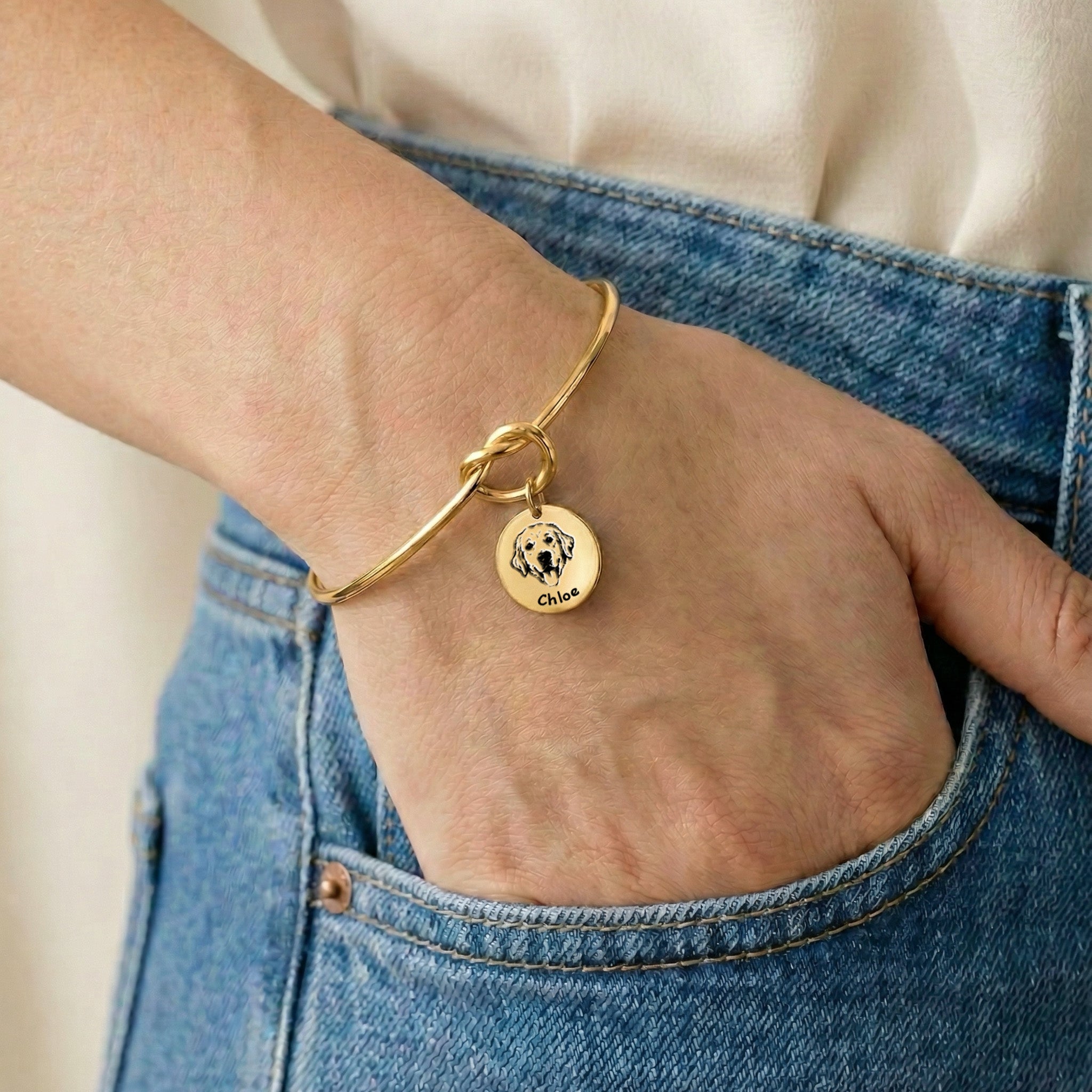Gold knot cuff bracelet with engraved dog portrait charm worn on a wrist, hand resting in a jeans pocket