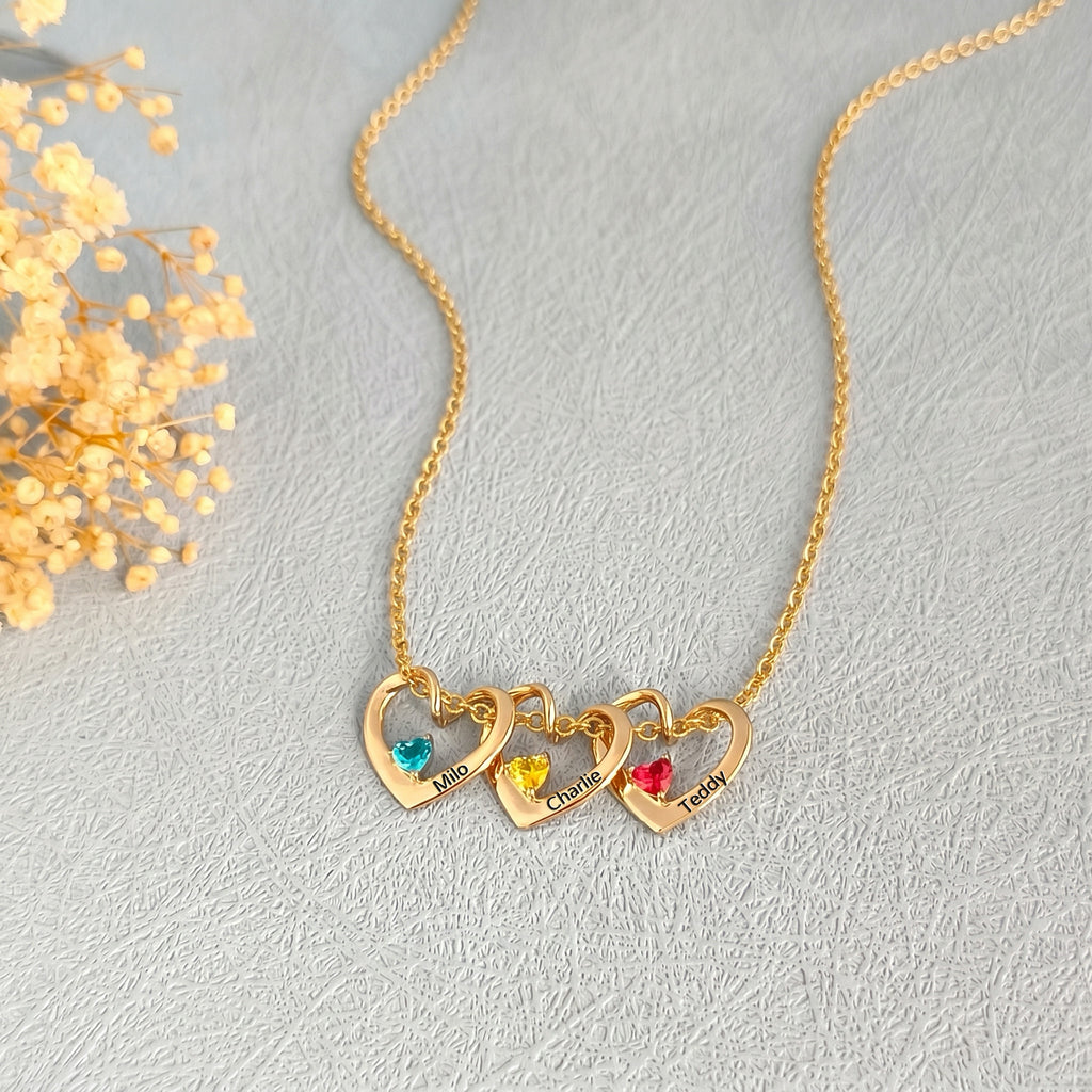 Three gold heart pendants with colorful birthstones and pet names on cream background