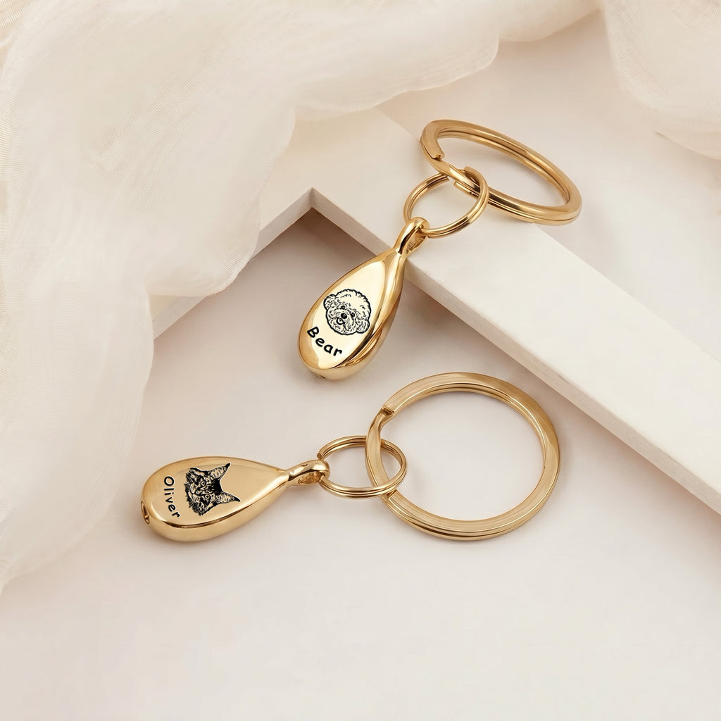 Pair of 18k gold-plated pet portrait ashes teardrop keychains arranged on a cream plinth under soft gauzy fabric &mdash; both pendants engraved with hand-drawn pet portraits and names