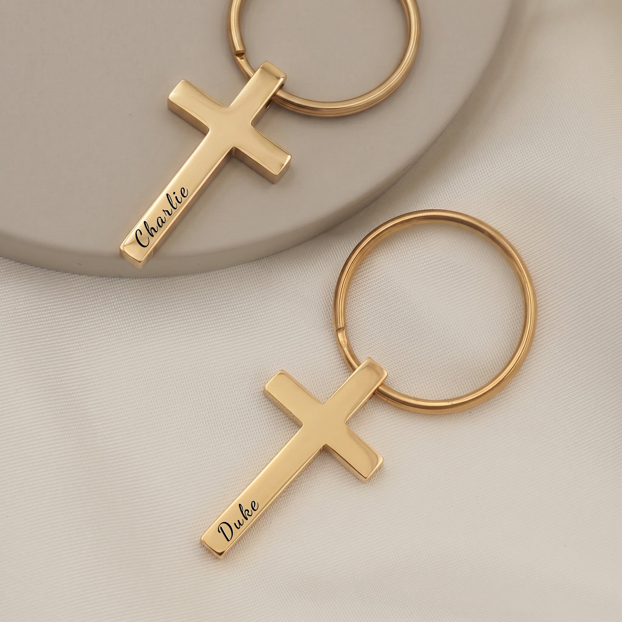 18k yellow gold pet cross keychain pair on ceramic tray with soft white fabric — front-face hand-drawn portrait + pet name engraving visible