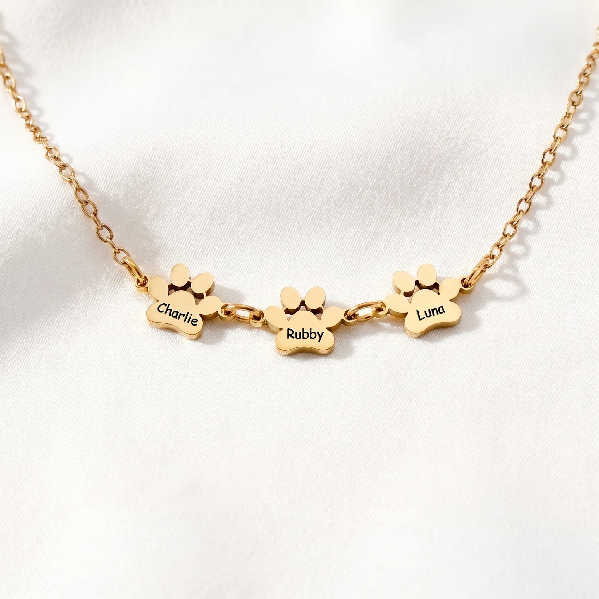 Three gold paw name charms linked along a bracelet chain, flat lay on white satin