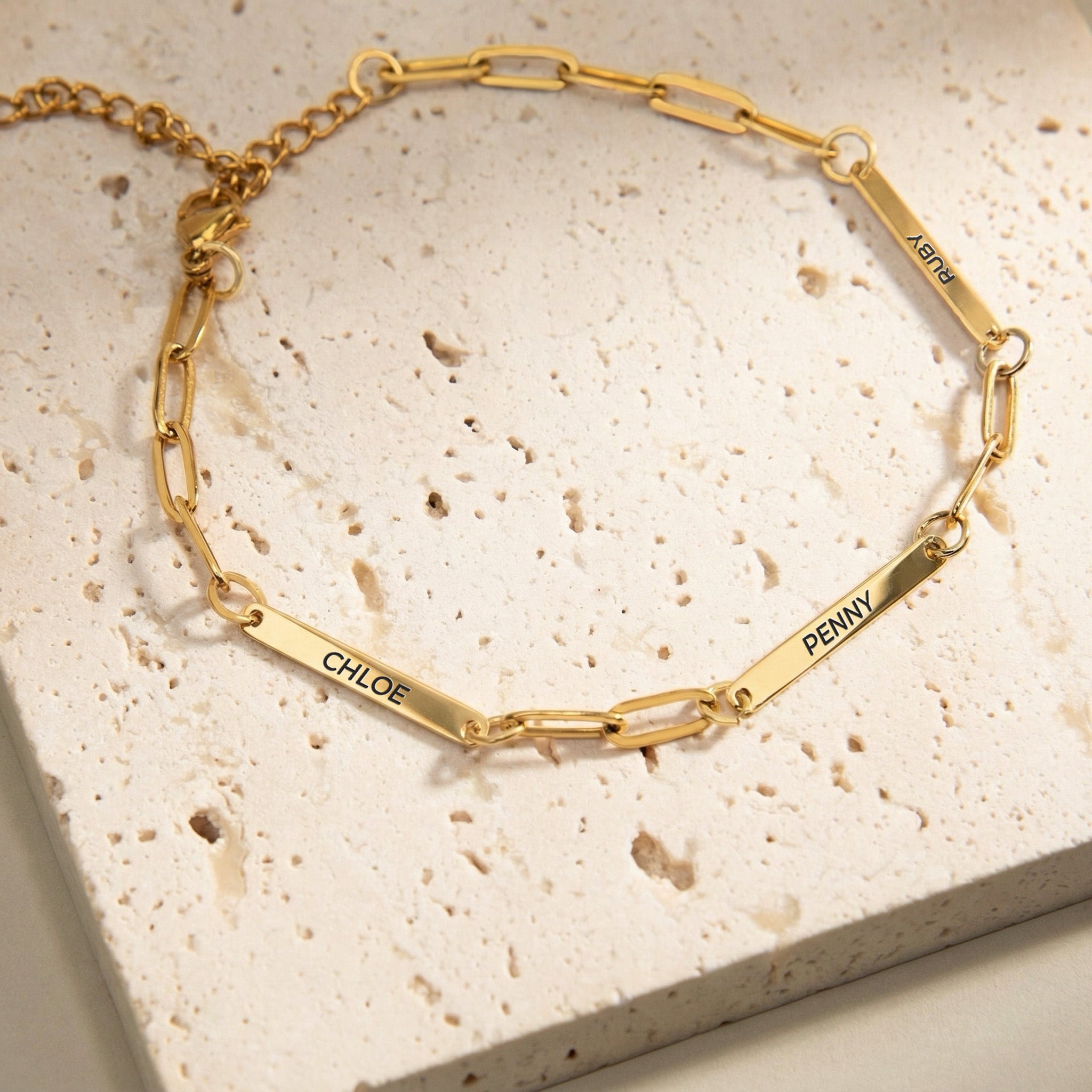 Gold paperclip chain pet name bracelet with three engraved name bars on a cream stone block