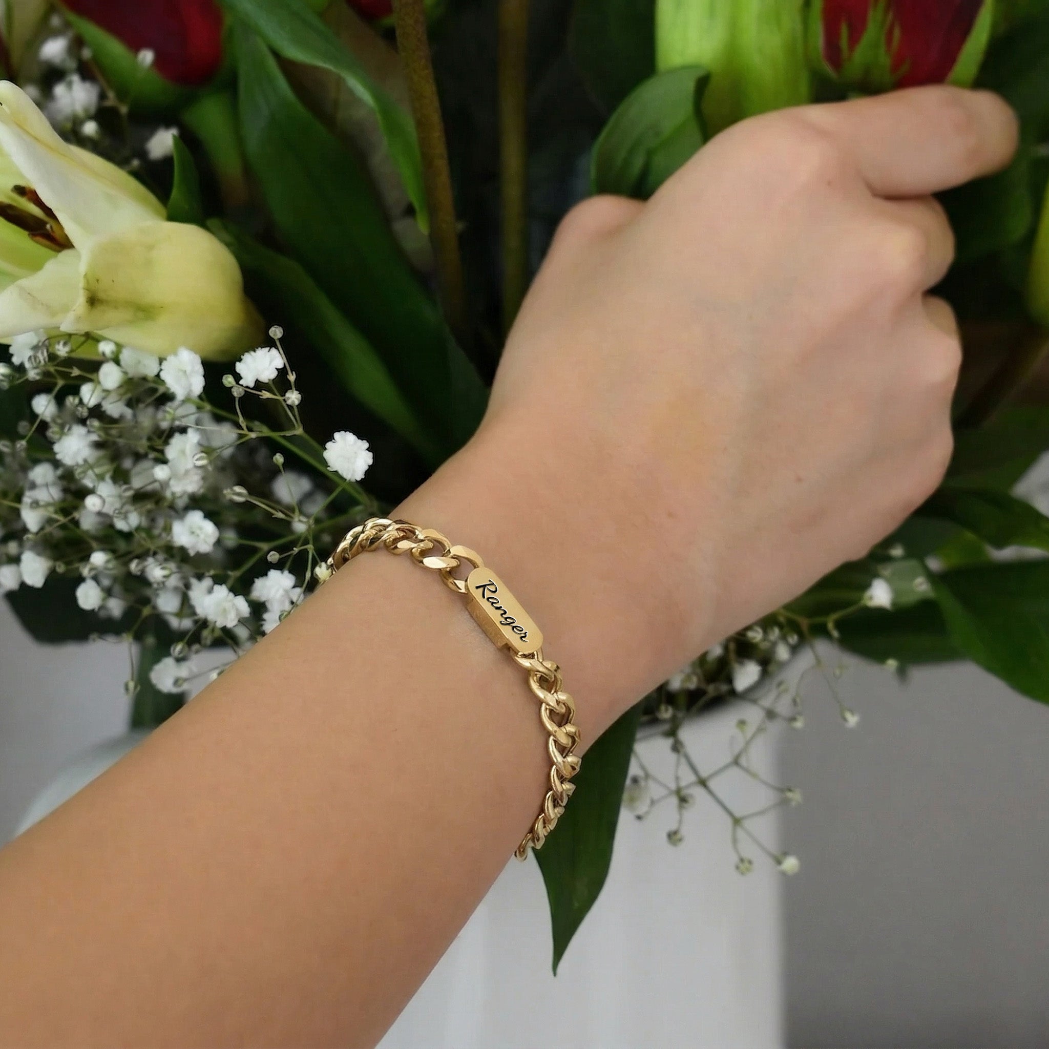 18K gold pet name bracelet with a script-engraved nameplate, worn on the wrist beside flowers