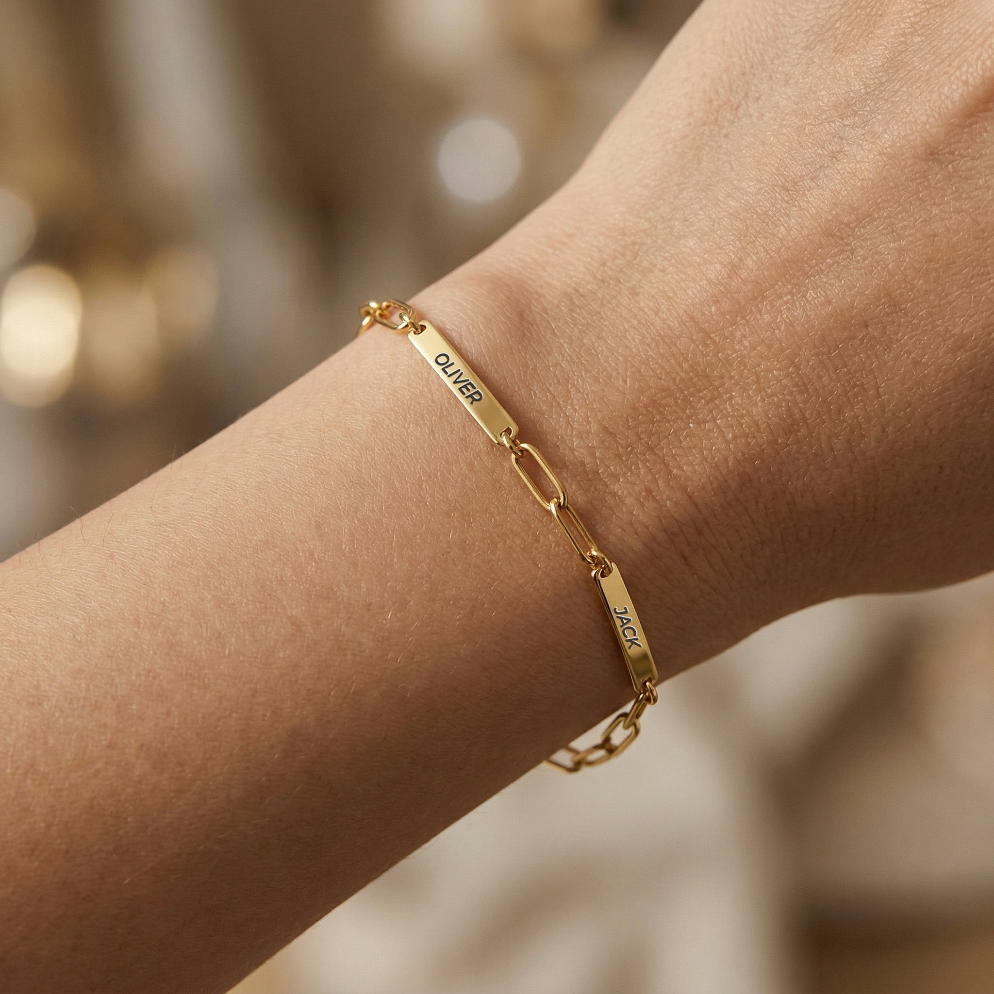 Gold paperclip chain pet name bracelet with engraved name bars worn on a wrist with soft bokeh