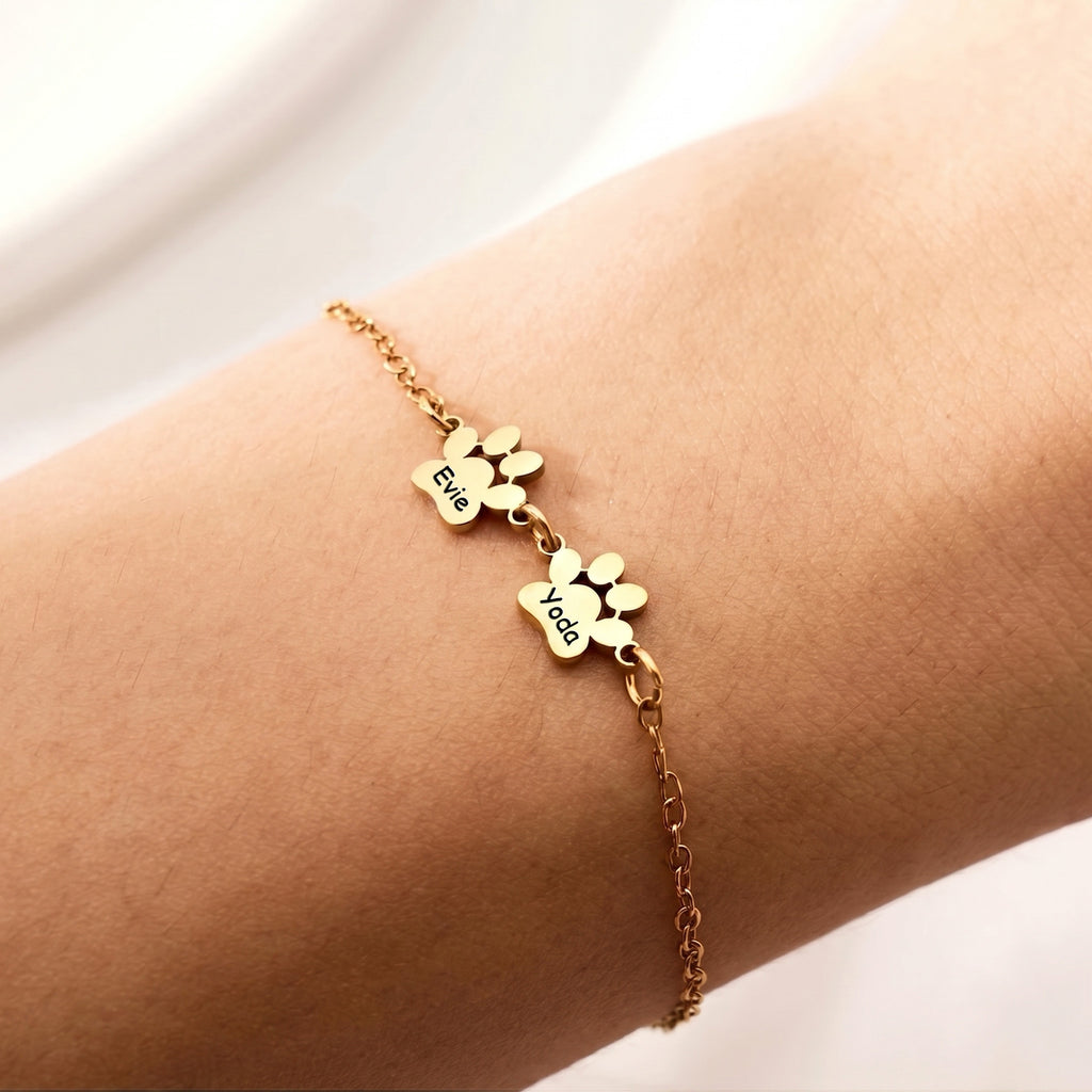 Gold paw print bracelet with two engraved name charms worn on the wrist in warm light