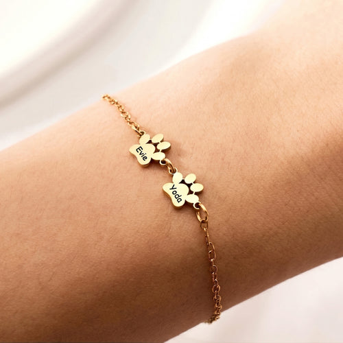 Gold paw print bracelet with two engraved name charms worn on the wrist in warm light