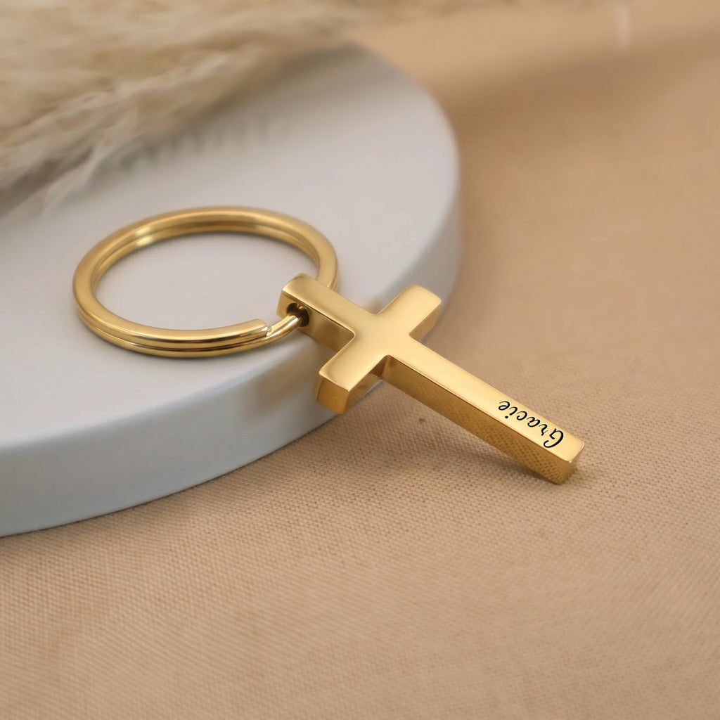 18k yellow gold pet cross keychain angled on a ceramic tray with beige fabric — full pendant and 25mm key ring visible in everyday-carry scene