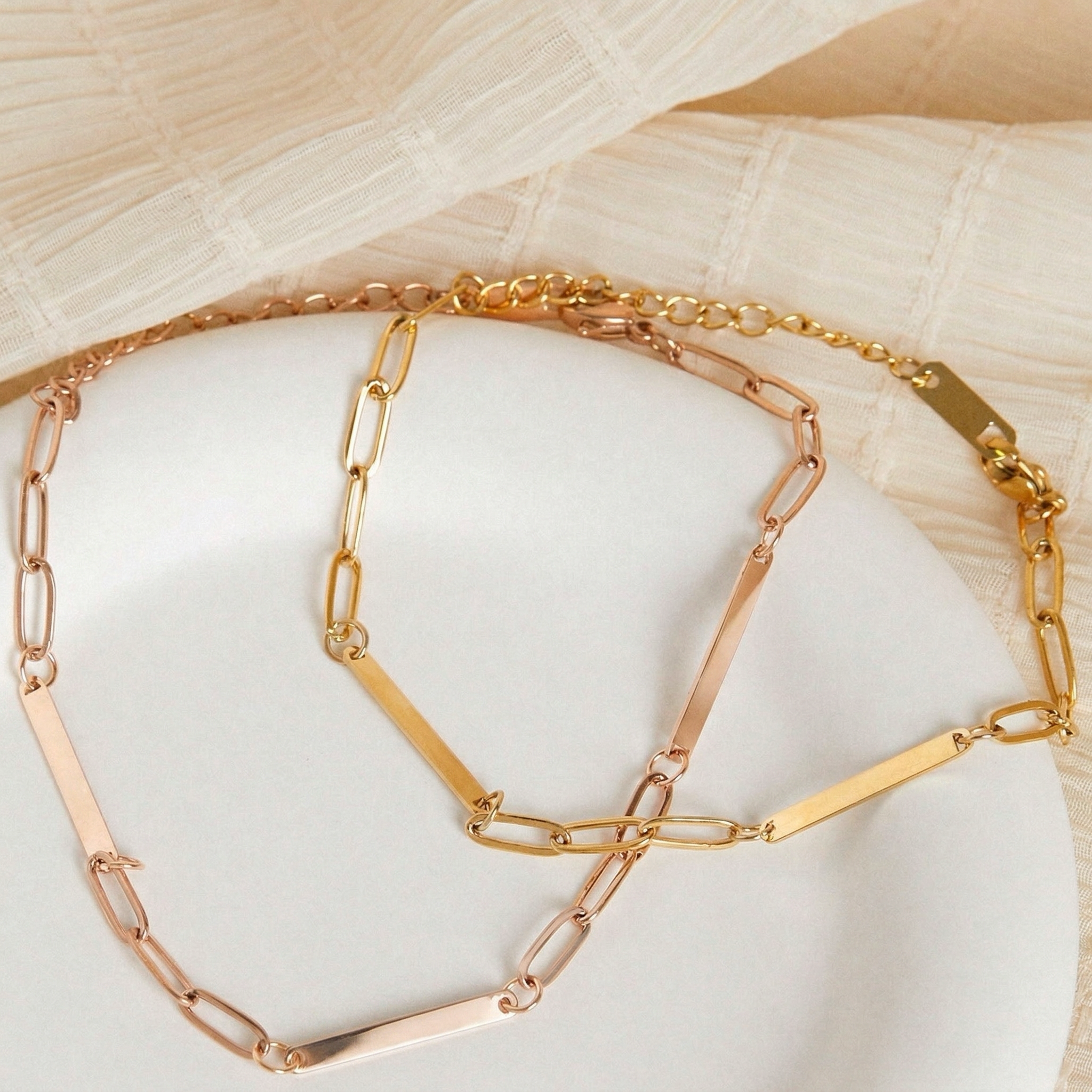 Gold and rose gold paperclip chain name bar bracelets draped on a white plate with cream fabric