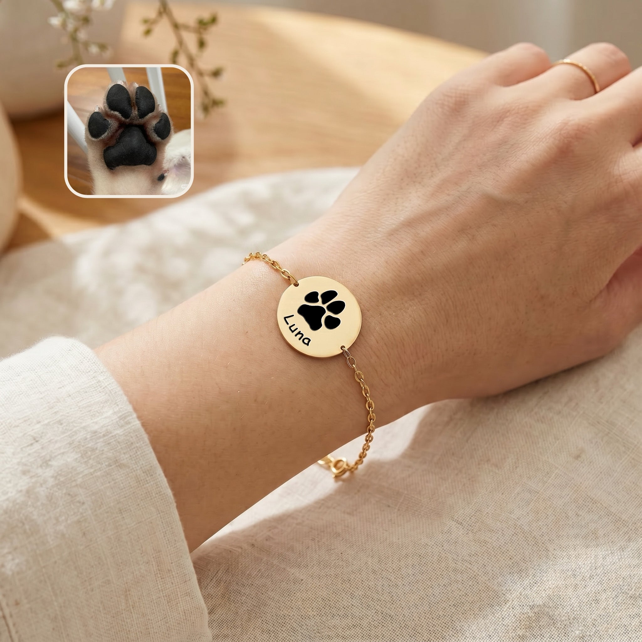 Gold-plated chain bracelet on a wrist showing paw print engraving and name on disc charm, linen background