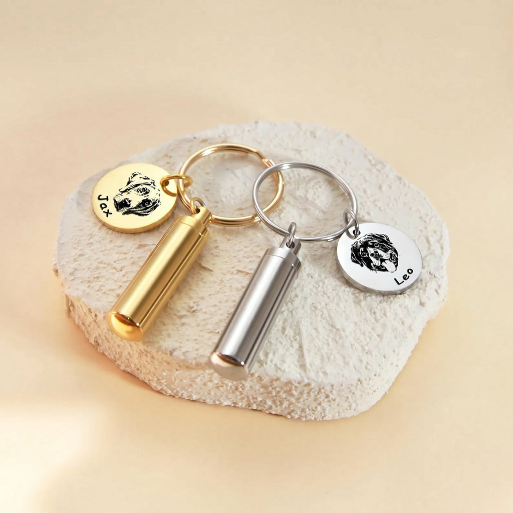 Pet portrait and ashes cylinder keychains shown in both finishes &mdash; 18k gold and rhodium silver paired on a textured stone surface
