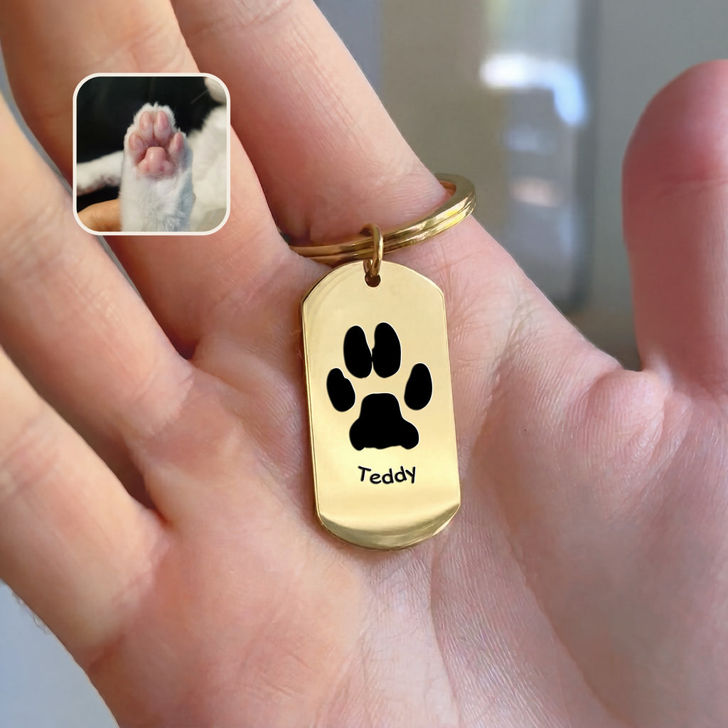 18K gold dog tag keychain with paw-print motif and pet name engraving, held in hand beside paw reference photo, warm indoor styled scene