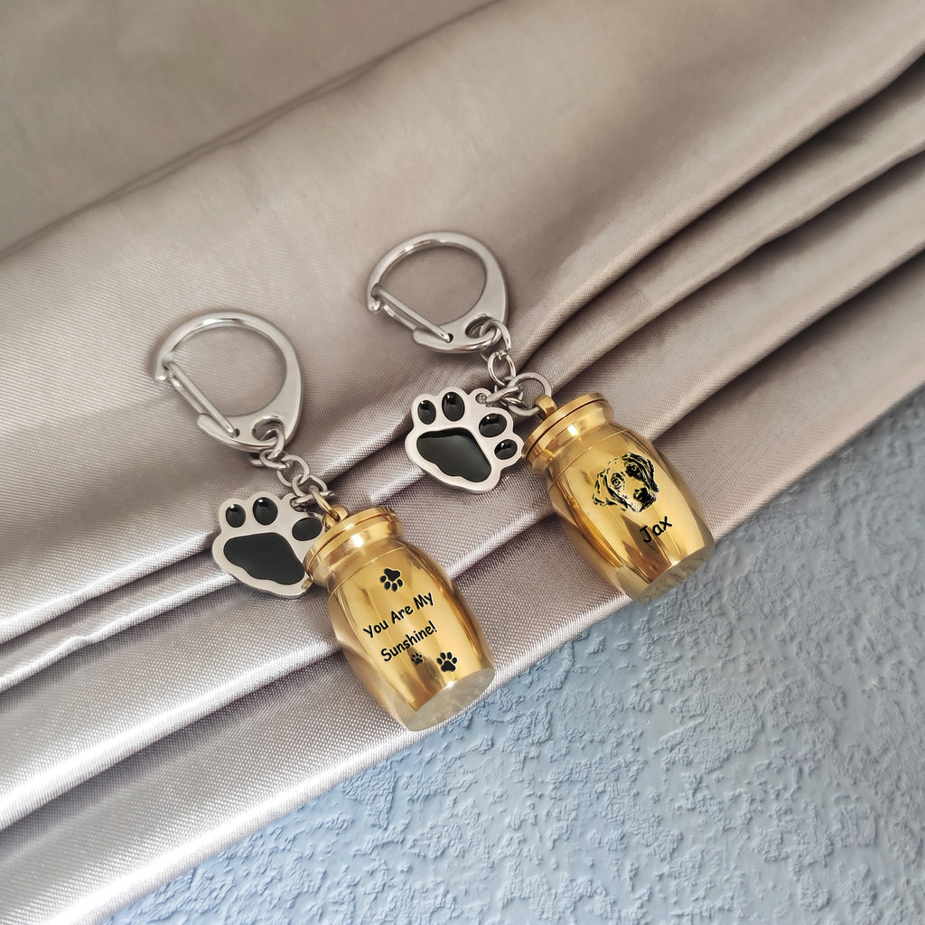 Two 18k yellow gold pet urn keychains arranged on draped satin fabric, one engraved with a verse and name, the other engraved with a hand-drawn portrait and name