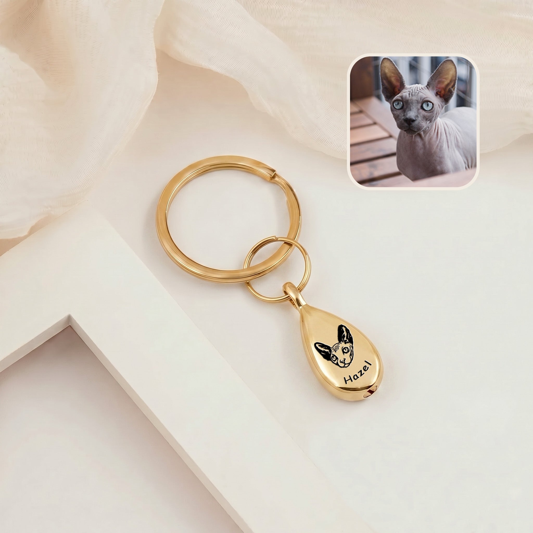 18k gold-plated pet portrait ashes teardrop keychain on a cream ledge with sheer fabric &mdash; engraved with a hand-drawn cat portrait and name, with a cat reference photo inset