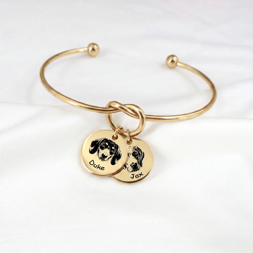 Gold knot cuff bracelet with two engraved pet portrait disc charms on a white background