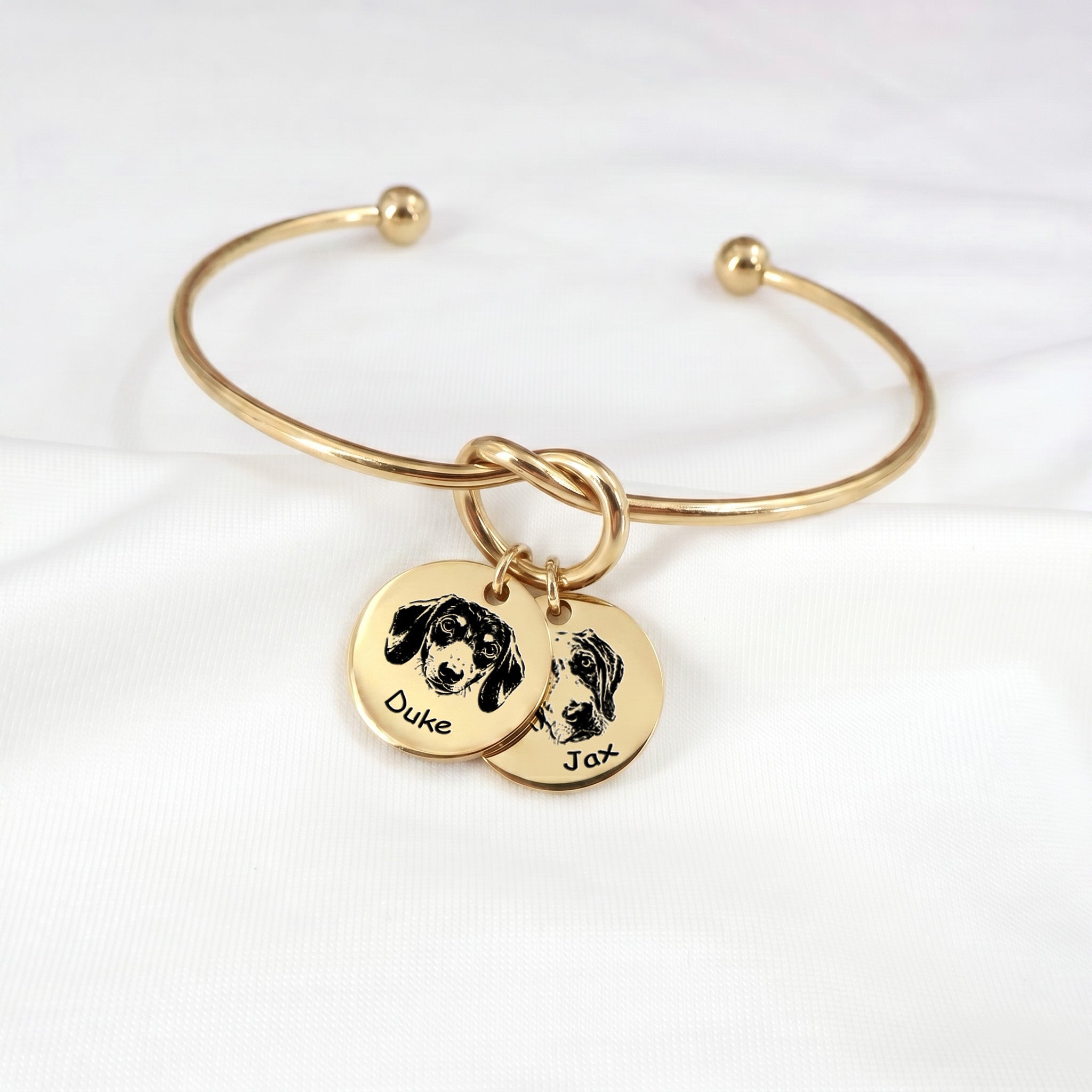 Gold knot cuff bracelet with two engraved pet portrait disc charms on a white background