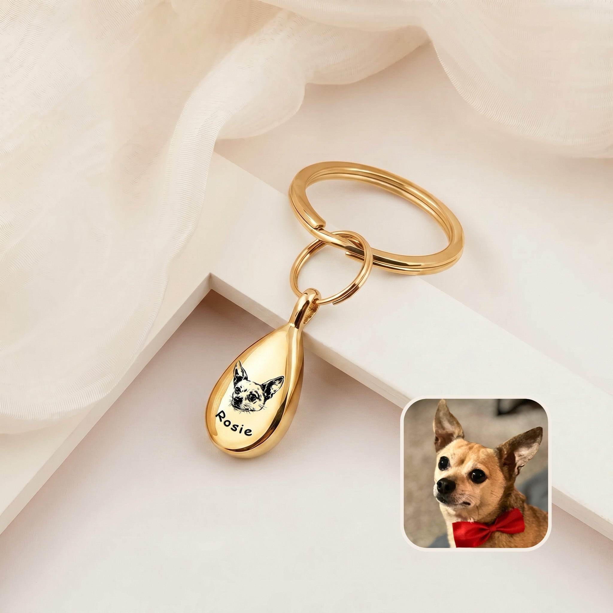 18k gold-plated pet portrait ashes teardrop keychain on a cream ledge with sheer fabric &mdash; engraved with a hand-drawn dog portrait and name, with a dog reference photo inset